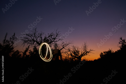 Silhouette of a man rotating light against trees and sunset sky background in long exposu photo