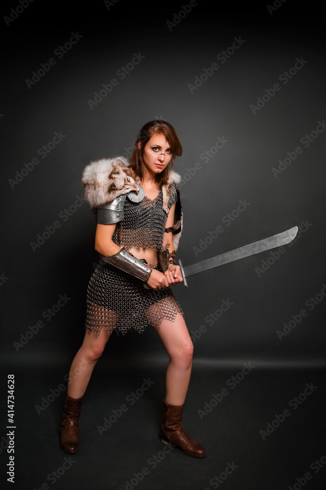Medieval woman warrior in chain mail armor with lamellar bracers and lamellar shoulder pads with polar fox fur on her shoulders stands with a sword in her hands against a dark background. Medieval woman warrior in chain mail armor with lamellar bracers and lamellar shoulder pads with polar fox fur on her shoulders stands with a sword in her hands against a dark background.