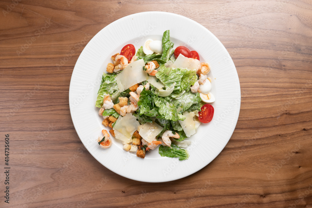 Caesar salad with prawns, lettuce, quail eggs, cherry tomatoes, croutons and parmesan cheese in a white ceramic plate on the wooden kitchen table. Caesar salad with prawns, lettuce, quail eggs, cherry tomatoes, croutons and parmesan cheese in a white ceramic plate on the wooden kitchen table.