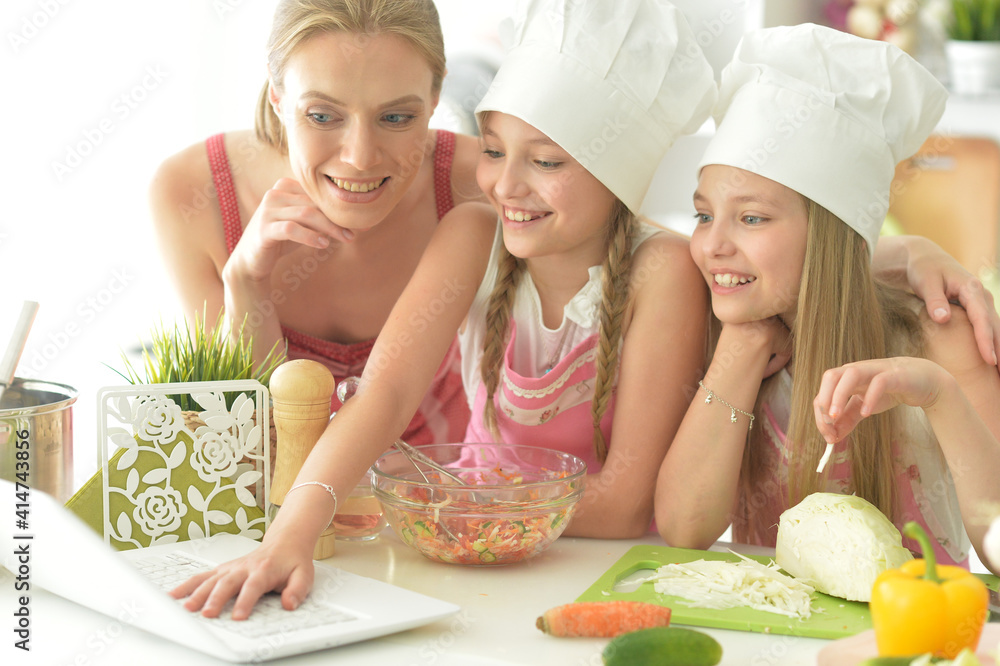 Cute girls with mother  preparing delicious fresh salad in kitchen looking at laptop