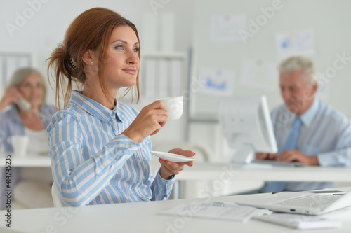 Beautiful young woman with cup in office, colleagues on bckground