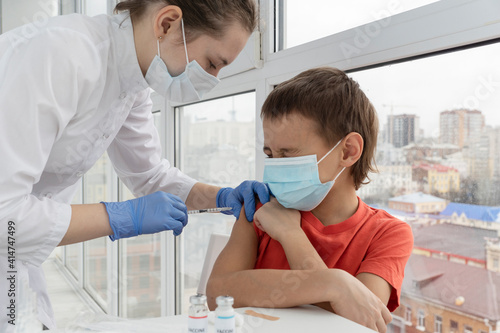 Boy in medical mask closed his eyes and as very afraid of vaccination. Young female doctor is making injection. Vaccination concept.