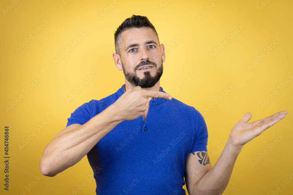 Handsome man with beard wearing blue polo shirt over yellow background surprised, showing and pointing something that is on her hand Handsome man with beard wearing blue polo shirt over yellow background surprised, showing and pointing something that is on her hand