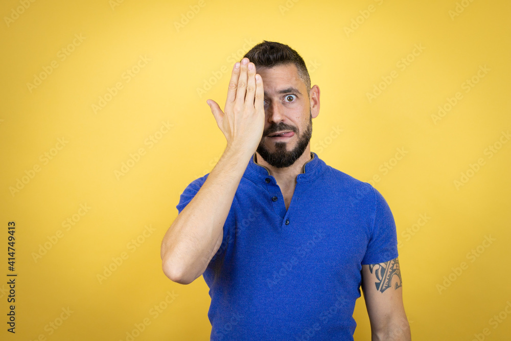 Handsome man with beard wearing blue polo shirt over yellow background covering one eye with hand, confident smile on face and surprise emotion. Handsome man with beard wearing blue polo shirt over yellow background covering one eye with hand, confident smile on face and surprise emotion.
