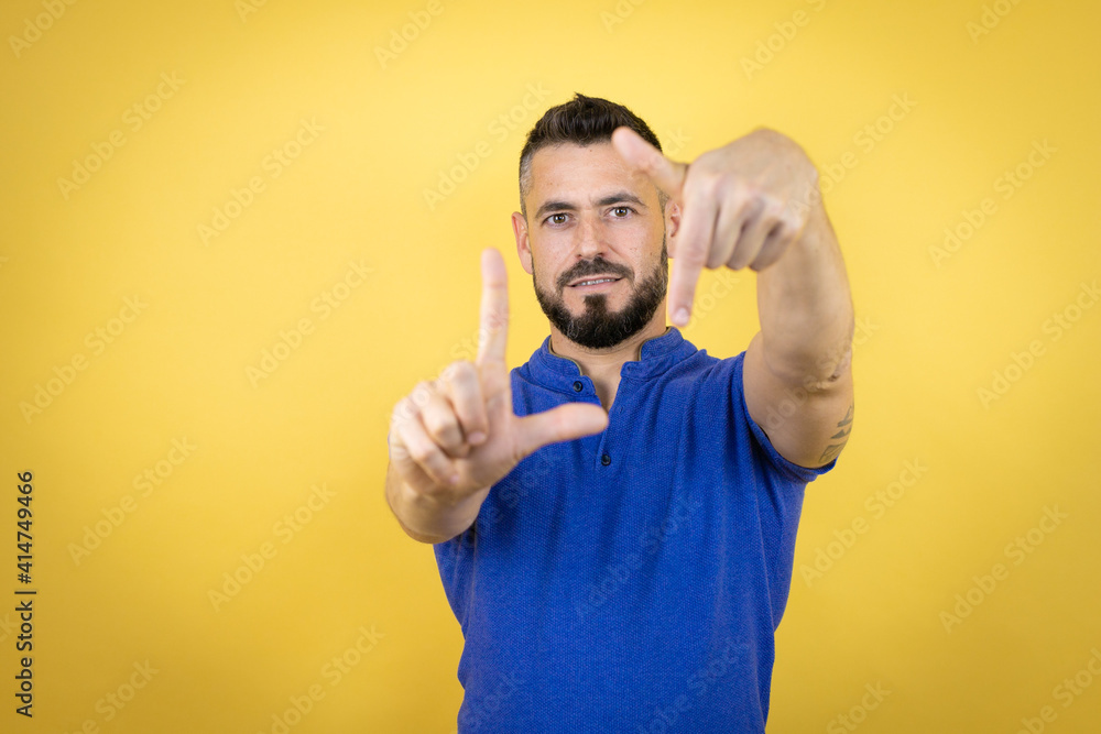 Handsome man with beard wearing blue polo shirt over yellow background smiling making frame with hands and fingers with happy face Handsome man with beard wearing blue polo shirt over yellow background smiling making frame with hands and fingers with happy face