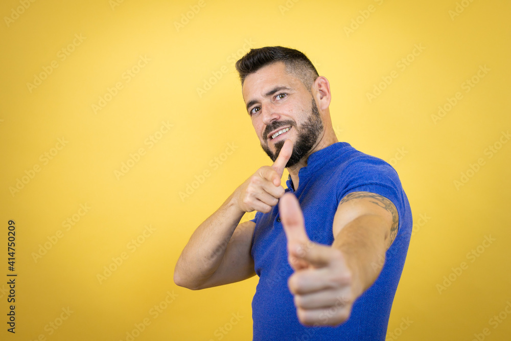 Handsome man with beard wearing blue polo shirt over yellow background pointing to you and the camera with fingers, smiling positive and cheerful Handsome man with beard wearing blue polo shirt over yellow background pointing to you and the camera with fingers, smiling positive and cheerful