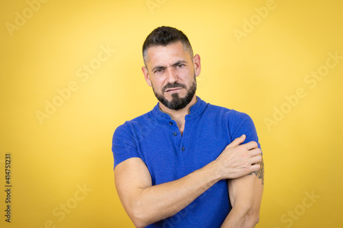 Handsome man with beard wearing blue polo shirt over yellow background with pain on his shoulder and a painful expression