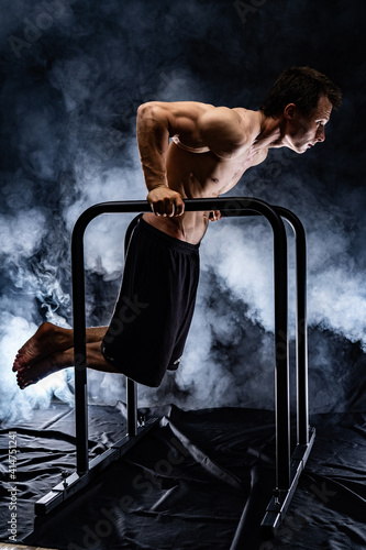 Muscular build man doing calisthenics on parallels bar indoor on black, smoked background. concept of motivation, desire and passion