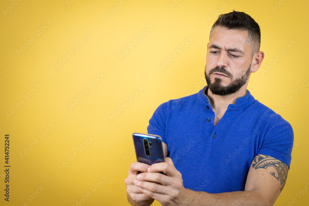 Handsome man with beard wearing blue polo shirt over yellow background chatting with his phone Handsome man with beard wearing blue polo shirt over yellow background chatting with his phone