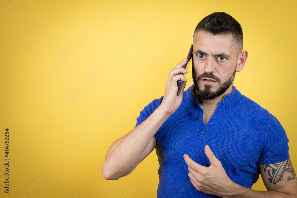 Handsome man with beard wearing blue polo shirt over yellow background talking on the phone with a worried expression Handsome man with beard wearing blue polo shirt over yellow background talking on the phone with a worried expression