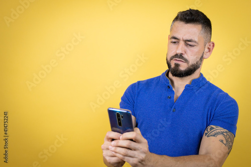 Handsome man with beard wearing blue polo shirt over yellow background chatting with his phone