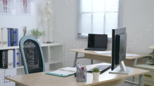 close up view office desk with a desktop, a notebook, a pen holder and a tiny pot plant in an empty doctor’s room on a sunny day.
