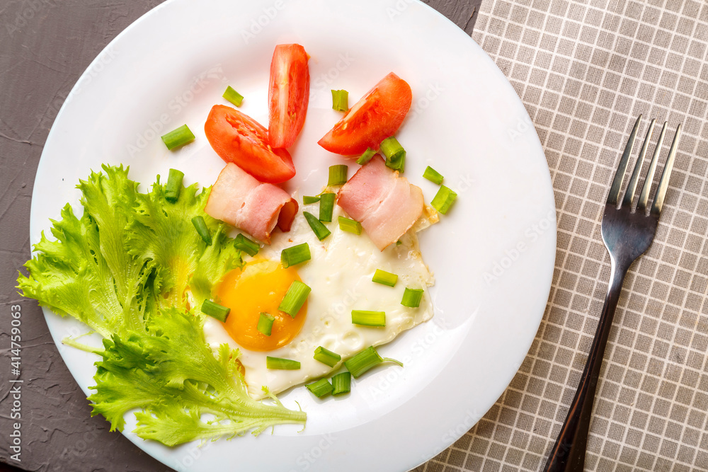 Scrambled eggs with bacon garnished with tomato and green onion on a gray concrete background on a napkin next to a fork. Scrambled eggs with bacon garnished with tomato and green onion on a gray concrete background on a napkin next to a fork.