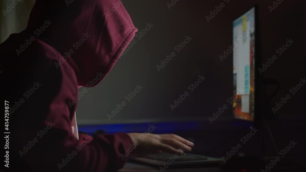 An unidentified man in the hood sits night for a computer monitor, password hacking, side view, cyber attack An unidentified man in the hood sits night for a computer monitor, password hacking, side view, cyber attack