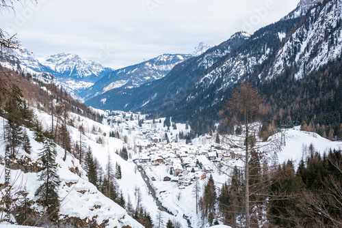 View form above of a town on the bank of a river running at the bottom of a forested mountain valley covered in snow in the European Alps on a cloudy winter day