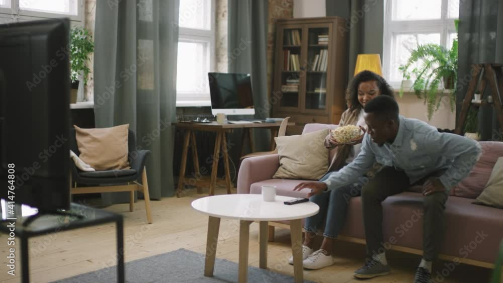 Panning slow-motion full shot of young happy afro couple sitting on couch in living room with popcorn, turning on TV and watching film together cuddling Panning slow-motion full shot of young happy afro couple sitting on couch in living room with popcorn, turning on TV and watching film together cuddling