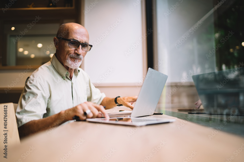 Aged freelancer in classic eyeglasses browsing received information on digital tablet sitting at desk with laptop computer, mature Caucaisan man 60s reading news and notifications on touch pad Aged freelancer in classic eyeglasses browsing received information on digital tablet sitting at desk with laptop computer, mature Caucaisan man 60s reading news and notifications on touch pad