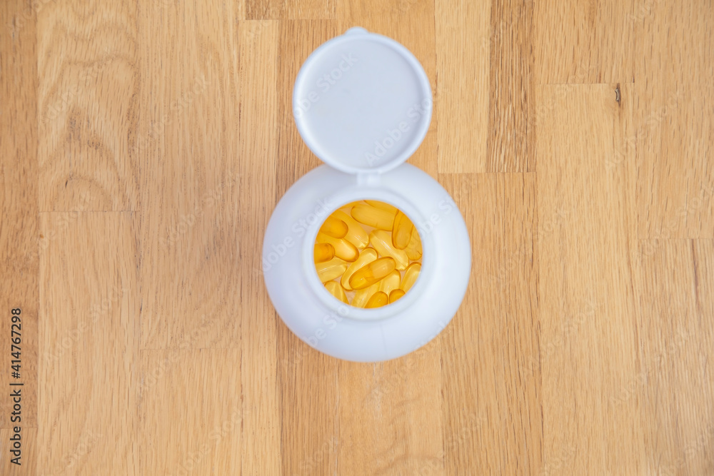 White jar with yellow transparent capsules of fish oil (omega-3, vitamin E) stands on a wooden table top view. Supplements and vitamins for sports and health in daily life White jar with yellow transparent capsules of fish oil (omega-3, vitamin E) stands on a wooden table top view. Supplements and vitamins for sports and health in daily life