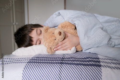 kid, a boy of 8 years old lies in bed, gently hugs a teddy bear with his hand, focus on the toy, concept of anxious sleep in schoolchildren, insomnia, night fears