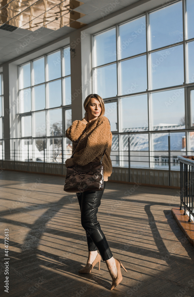 Fototapeta premium A stylish girl in a brown sweater, black pants with a large women's bag poses against the background of large windows.