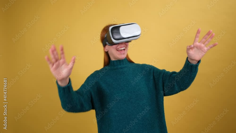 Amazed young woman using VR app headset helmet to play simulation game, drawing. Watching virtual reality 3d 360 video. Isolated on yellow background in studio. Amazed young woman using VR app headset helmet to play simulation game, drawing. Watching virtual reality 3d 360 video. Isolated on yellow background in studio.
