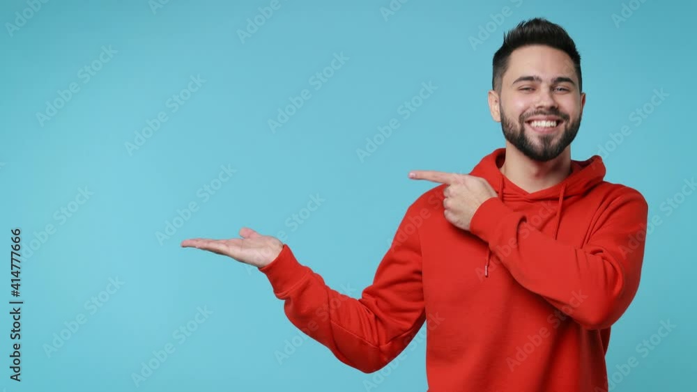 Cheerful funny young man 20s years old in casual red streetwear hoodie isolated on blue background studio. People lifestyle concept. Pointing index finger on empty palm showing thumb up like gesture Cheerful funny young man 20s years old in casual red streetwear hoodie isolated on blue background studio. People lifestyle concept. Pointing index finger on empty palm showing thumb up like gesture