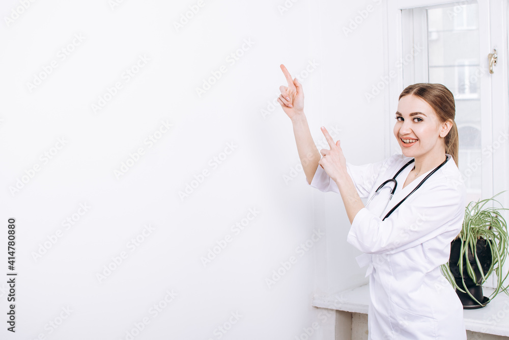 Smiling happy medical doctor woman points with finger on white background near window. Advertising of a clinic, pharmacy, medicine, vaccination.