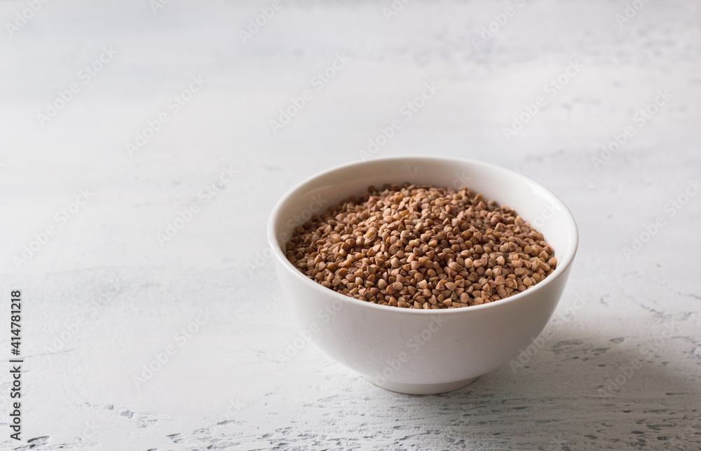 Raw buckwheat in a white ceramic bowl on a light gray textured backgroundfree space
