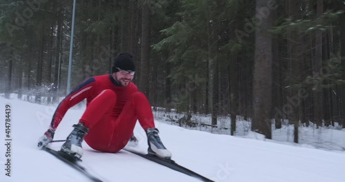 Skier falls in a funny way. Young man in red ski suit glides downhill on the cross country ski track and falls on his buttocks