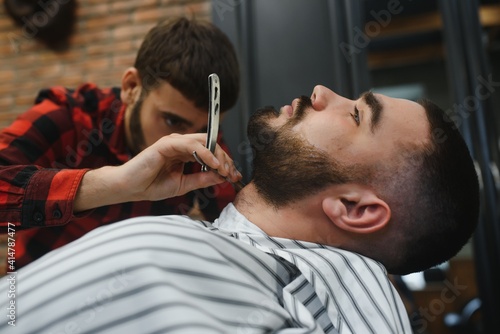 Serious Bearded Man Getting Beard Haircut With A Straight Razor By Barber While Sitting In Chair At Barbershop. Barbershop Theme
