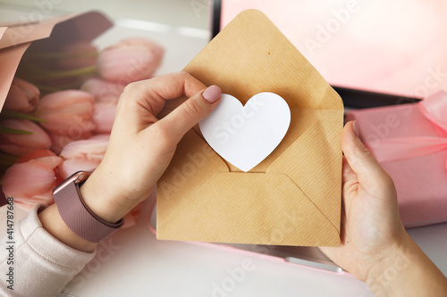 Woman's hands holding a letter in craft envelope. Pink background, valentine's day concept. Tulips flower and pink gift box in background. Womens home desk