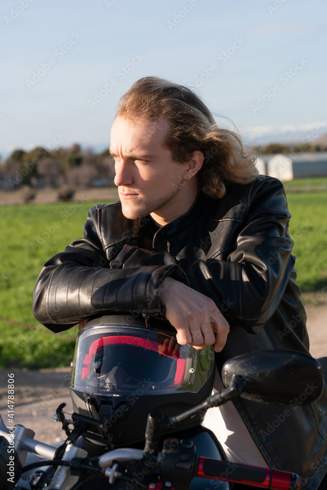 Caucasian white man leaning on a black motorcycle biker helmet with sane jacket