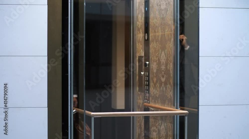 Personable bearded male in business suit talking on smartphone while entering elevator car. Confident businessman going down in transparent lift cabin while leaving hotel photo