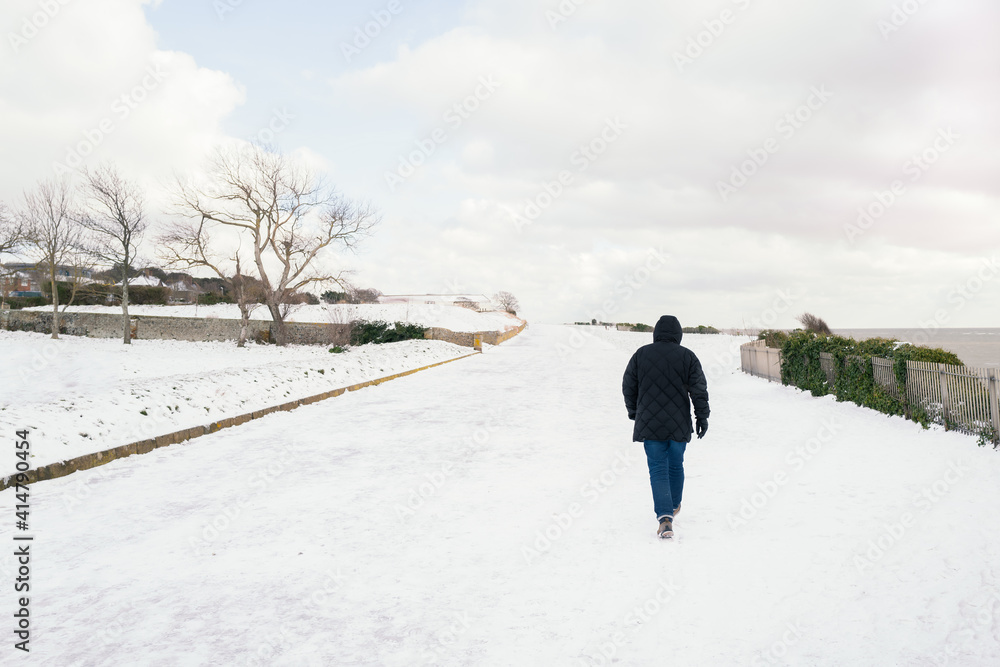 A man walks along an empty snow covered promenade during the day. The clouds are white. There are trees on one side and the sea on the other.