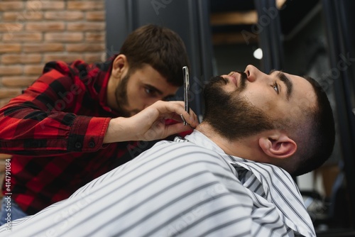 Serious Bearded Man Getting Beard Haircut With A Straight Razor By Barber While Sitting In Chair At Barbershop. Barbershop Theme