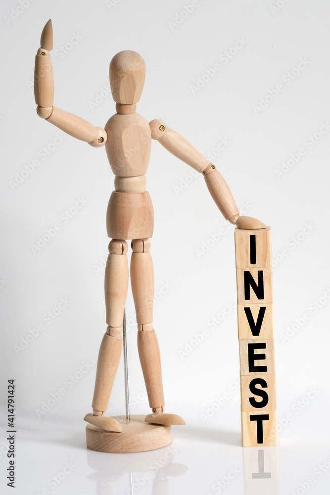 The wooden man raised his hand, and under his left hand he has cubes with the inscription - INVEST The wooden man raised his hand, and under his left hand he has cubes with the inscription - INVEST