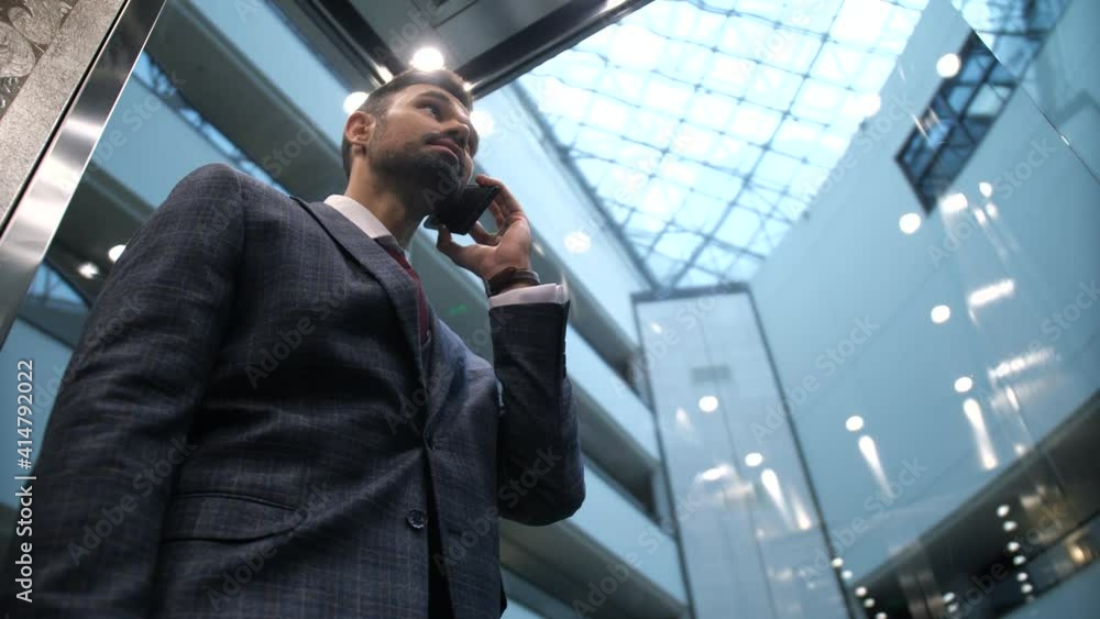 Low angle view of smiling bearded businessman talking on smartphone while going down in transparent lift of luxury hotel. Handsome enterpreneur in business suit during conversation in hotel elevator Low angle view of smiling bearded businessman talking on smartphone while going down in transparent lift of luxury hotel. Handsome enterpreneur in business suit during conversation in hotel elevator