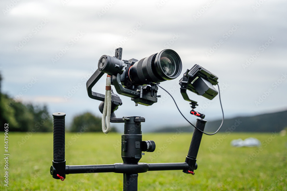 Camera in a professional video recording set planted on the ground, on top of a gimbal / stabilizer with included screen. Green background in the middle of mountains.