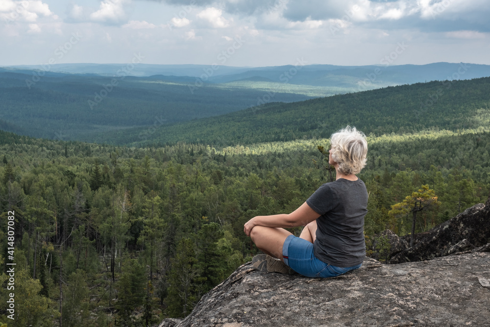 A middle aged woman practices yoga and meditation on a mountain top with a stunning view of a forested, hilly valley.