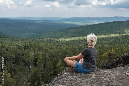 A middle aged woman practices yoga and meditation on a mountain top with a stunning view of a forested, hilly valley.