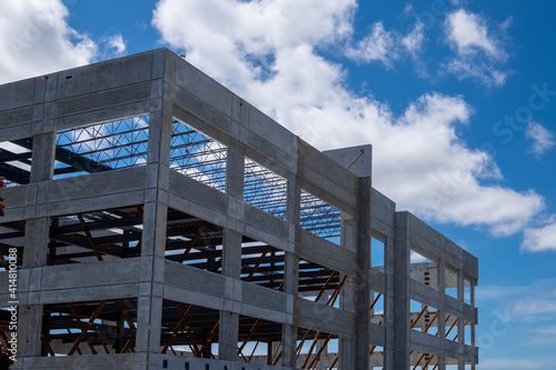A large multiple storey precast grey concrete building with steel beams against a blue sky. The industrial structure is the corner of a skyscraper building with prefabricated engineering formwork. © Dolores  Harvey