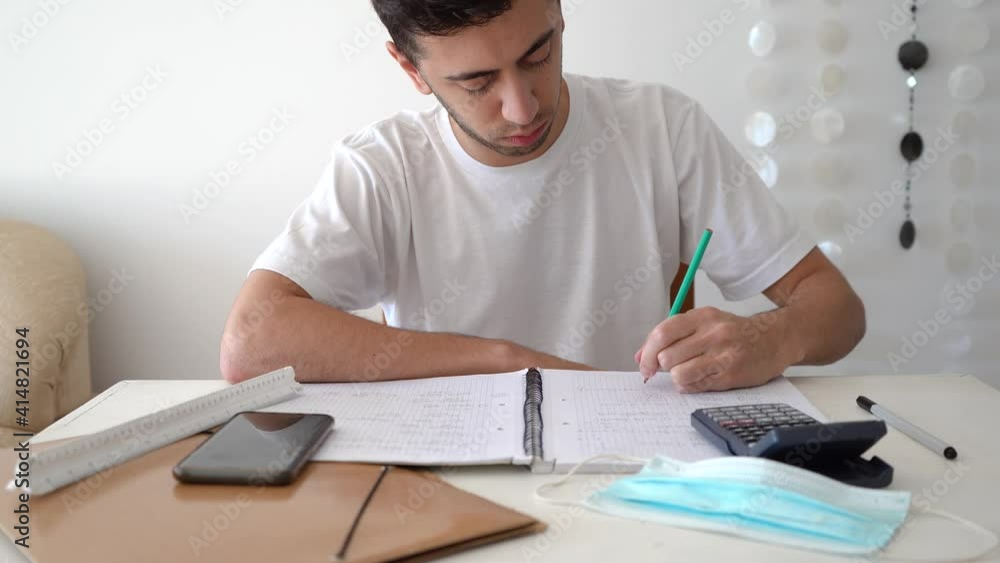 Male Student Writing Mathematical Formulas And Solutions On A Notebook With Calculator, Smartphone, And Medical Mask On The Table. medium shot Male Student Writing Mathematical Formulas And Solutions On A Notebook With Calculator, Smartphone, And Medical Mask On The Table. medium shot