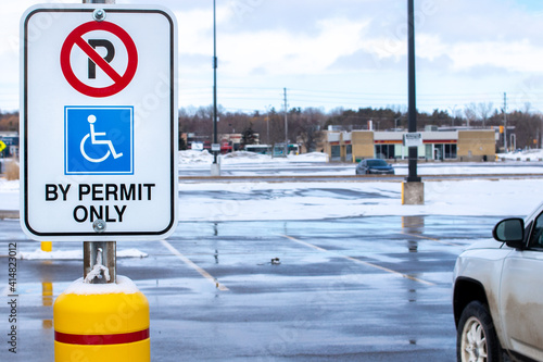 No parking, by permit only sign with the international access symbol displayed on a rectangular vertical sign in a mucky and snowy parking lot in the middle of winter in Canada.
