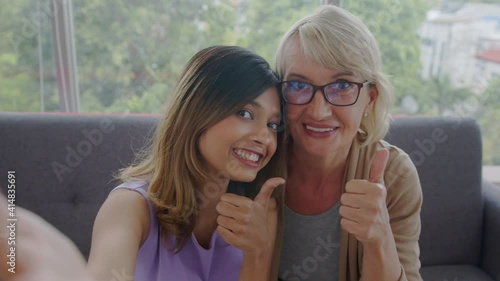Medium shot - Adopted Asian teen and her Caucasian stepmother enjoy talking a selfie together. Free-time and love sharing of biracial family. 