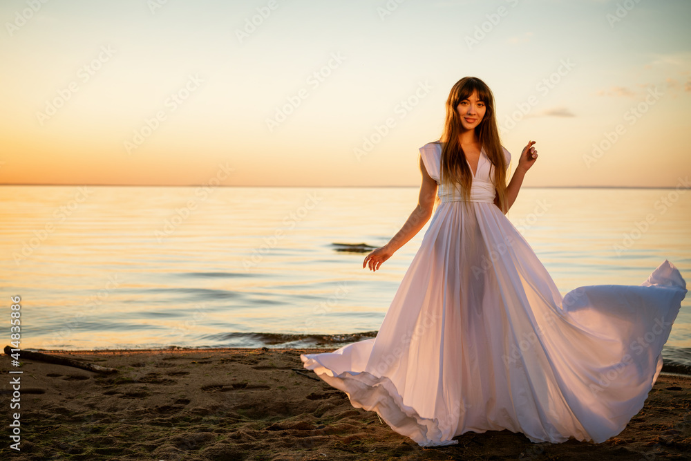 Beautiful happy young woman in a white long dress on the seashore at sunset stands