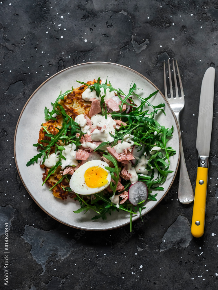 Potato latkes and salad with arugula, canned tuna, boiled egg, yogurt, mustard dressing on a dark background, top view Potato latkes and salad with arugula, canned tuna, boiled egg, yogurt, mustard dressing on a dark background, top view
