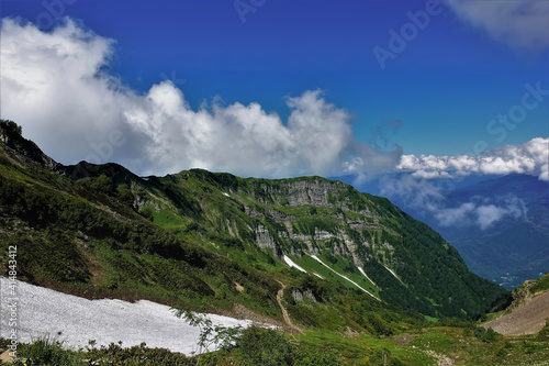 Caucasian mountain range against the background of blue sky and clouds. The rocky slopes of the mountains are covered with green vegetation. In the valley there are areas of melted snow. Summer day.