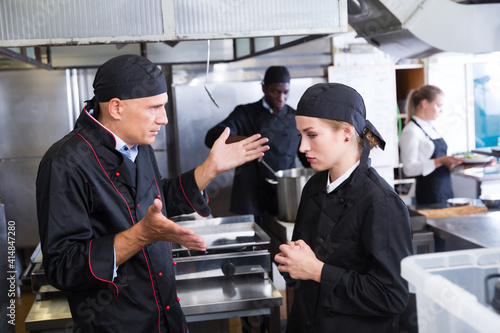 Enraged chef expressing dissatisfaction with work of frustrated girl in restaurant kitchen photo