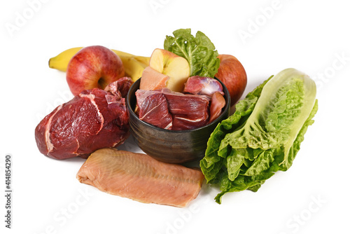 Dog bowl filled with biologically appropriate raw food containing meat chunks, fish, fruits and vegetables surrounded by ingredients on white background photo