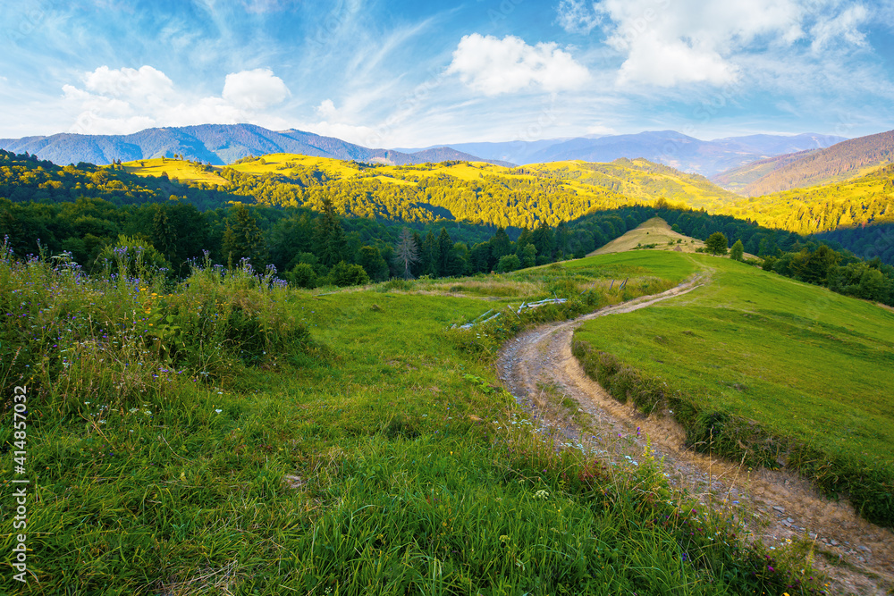 rural landscape in mountains at summer sunrise. country road through grassy pasture winding down in to the distant valley. clouds on the blue sky above the ridge in the distance